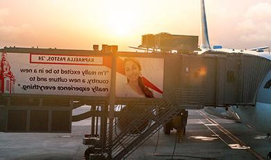 一架飞机停在登机口的图形，在登机梯上引用了拉斐尔·帕斯托斯(Raphaella Pastos)的一句话:“我真的很兴奋来到一个新的国家，一种新的文化，真正体验一切。”